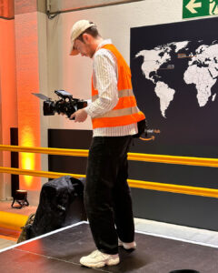 Mann mit Kappe und Sicherheitsweste filmt mit einer Kamera vor einer Weltkarte