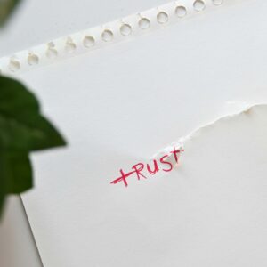 Ein weißes Blatt Papier mit einer roten handgeschriebenen Aufschrift 'TRUST', das in der Mitte zerrissen ist. Die Risskante verläuft durch das Wort, sodass es teilweise zerstört erscheint. Im Vordergrund ist ein unscharfes grünes Blatt einer Pflanze zu sehen.