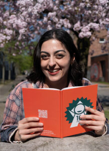 Eine junge Frau mit dunklen Haaren sitzt draußen vor einem blühenden Kirschbaum und hält ein orangefarbenes Notizbuch mit dem Logo von Wake up Communications in den Händen. Sie lacht herzlich in die Kamera. Auf dem Notizbuch ist ein QR-Code sowie der Text „Wake up Communications – Agentur für PR & Social Media“ zu sehen.